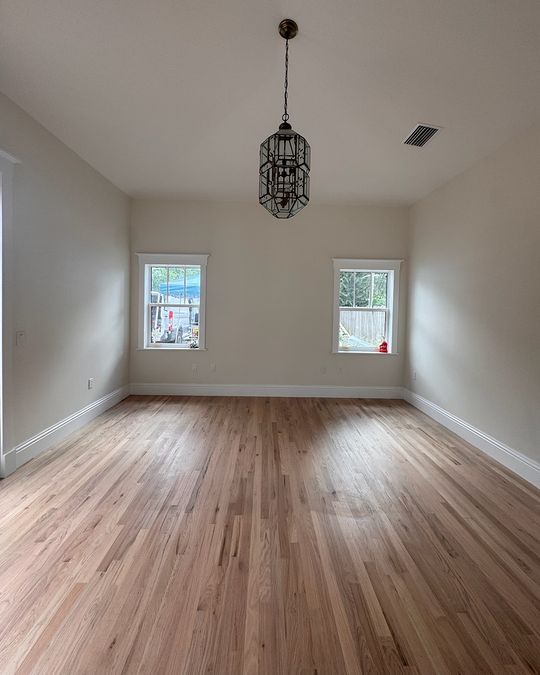 An empty bedroom with a natural oak hardwood floor and two windows.