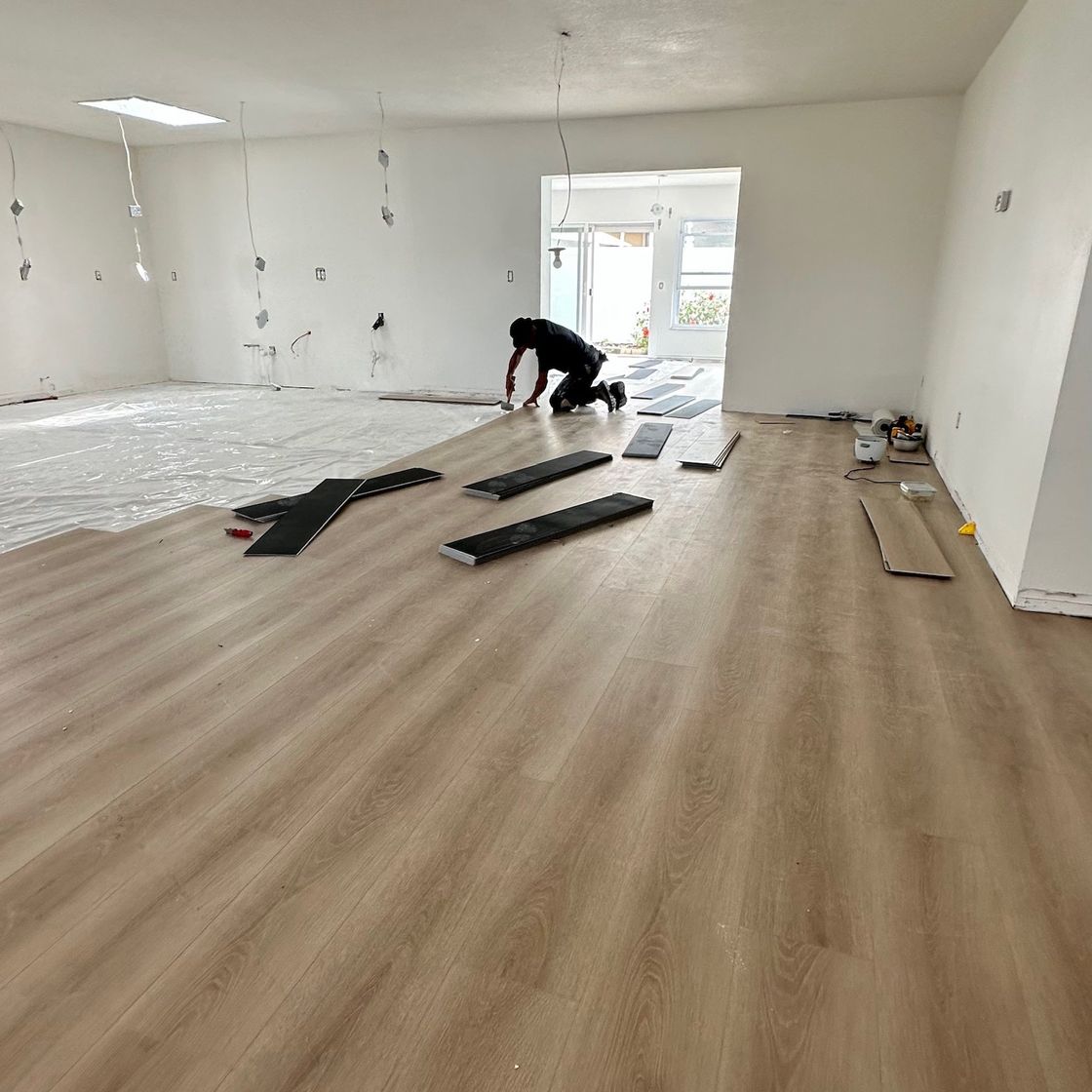 An installer kneeling to fit new wood-look plank flooring in a large open room, with the finished floor in the foreground.