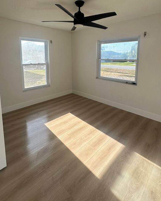 A bedroom with sunlight across a light wood-look plank floor and white baseboards.