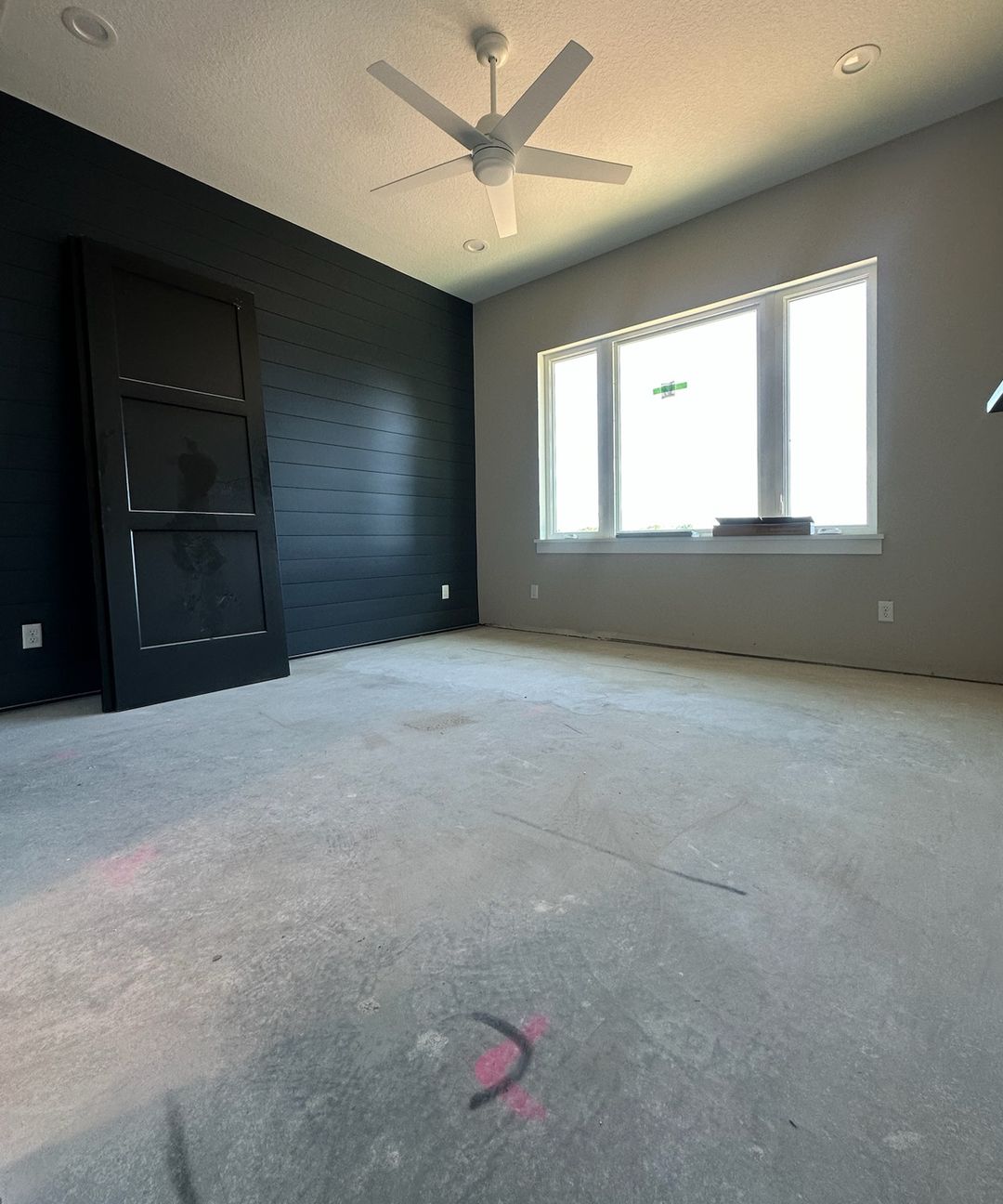A bedroom on day one: a bare concrete slab, a dark paneled wall and a wide window.